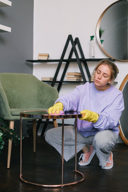 Close-up of a green upholstered armchair with a tufted backrest and a smooth seat cushion, being cleaned using a handheld vacuum cleaner. The vacuum hose is pressed against the fabric to remove dust a