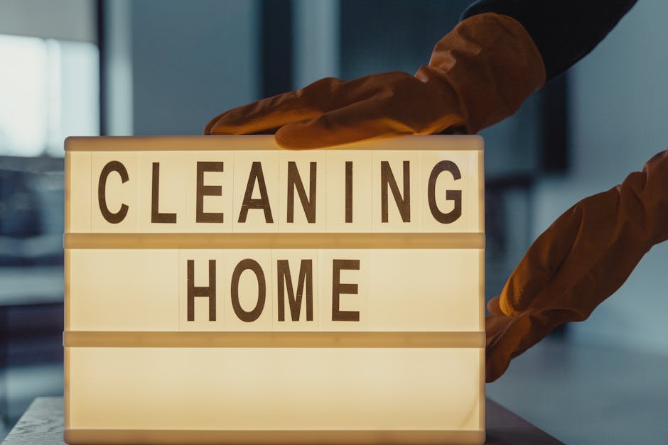 A person wearing orange cleaning gloves is adjusting a backlit sign with black letters spelling 'CLEANING HOME' on a table, set against a blurred background of an indoor space with natural light. The 