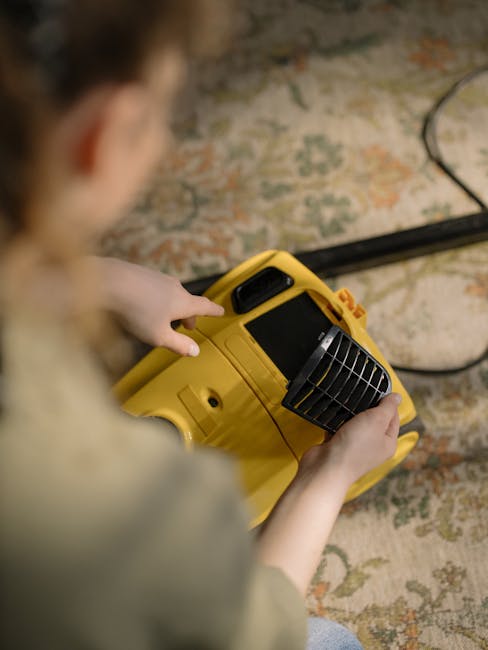 A person kneeling on a patterned carpet in a domestic setting, loading a portable yellow and black carpet cleaning machine with a transparent water tank and a flexible hose. The room features warm lig